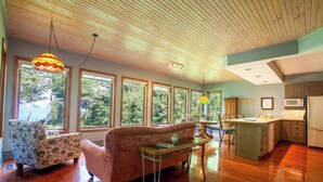 A spacious living room with large windows, a wooden ceiling, and a kitchen area.