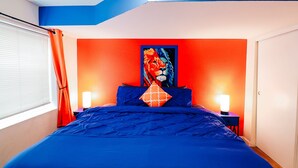 Ett sovrum med ett blått sängöverkast, orangea gardiner, en röd accentvägg och en tavla som föreställer ett lejon.