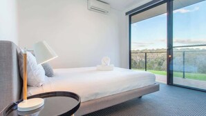 A modern hotel room with a bed, a bedside table, a lamp, and a large window offering a view of the outdoors.