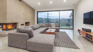 A modern living room with a large sectional sofa, a fireplace, a flat-screen TV, and a large window offering a view of the outdoors.