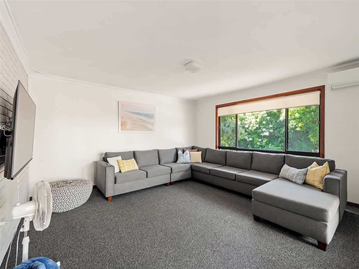 Un salon moderne avec un canapé d’angle gris, un petit pouf, un ventilateur sur pied et une grande fenêtre donnant sur un paysage verdoyant.
