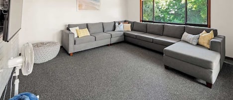 Un salon moderne avec un canapé d’angle gris, un petit pouf, un ventilateur sur pied et une grande fenêtre donnant sur un paysage verdoyant.