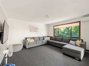 A modern living room with a grey sectional sofa, a small ottoman, a standing fan, and a large window offering a view of greenery.