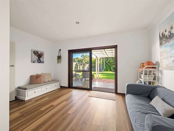 A spacious living room with a sliding glass door leading to a backyard, a grey sofa, and a wooden bench with cushions.