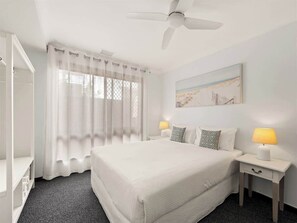 A hotel room with a bed, two bedside tables, a ceiling fan, and a window with sheer curtains.