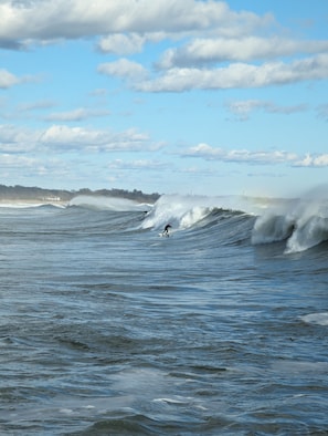A surfer riding a large wave in the ocean with a clear sky and distant shoreline. - Entire House:14 Guests, 7 BRs, 10 Beds, 5 Full Baths, Outdoor Shower, Beach Gear (Narragansett Pier)