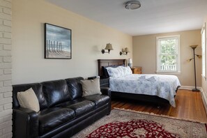 A bedroom with a bed, a black leather sofa, a red and white patterned rug, a painting of a beach, and a window with blinds. - Entire House:14 Guests, 7 BRs, 10 Beds, 5 Full Baths, Outdoor Shower, Beach Gear (Narragansett Pier)