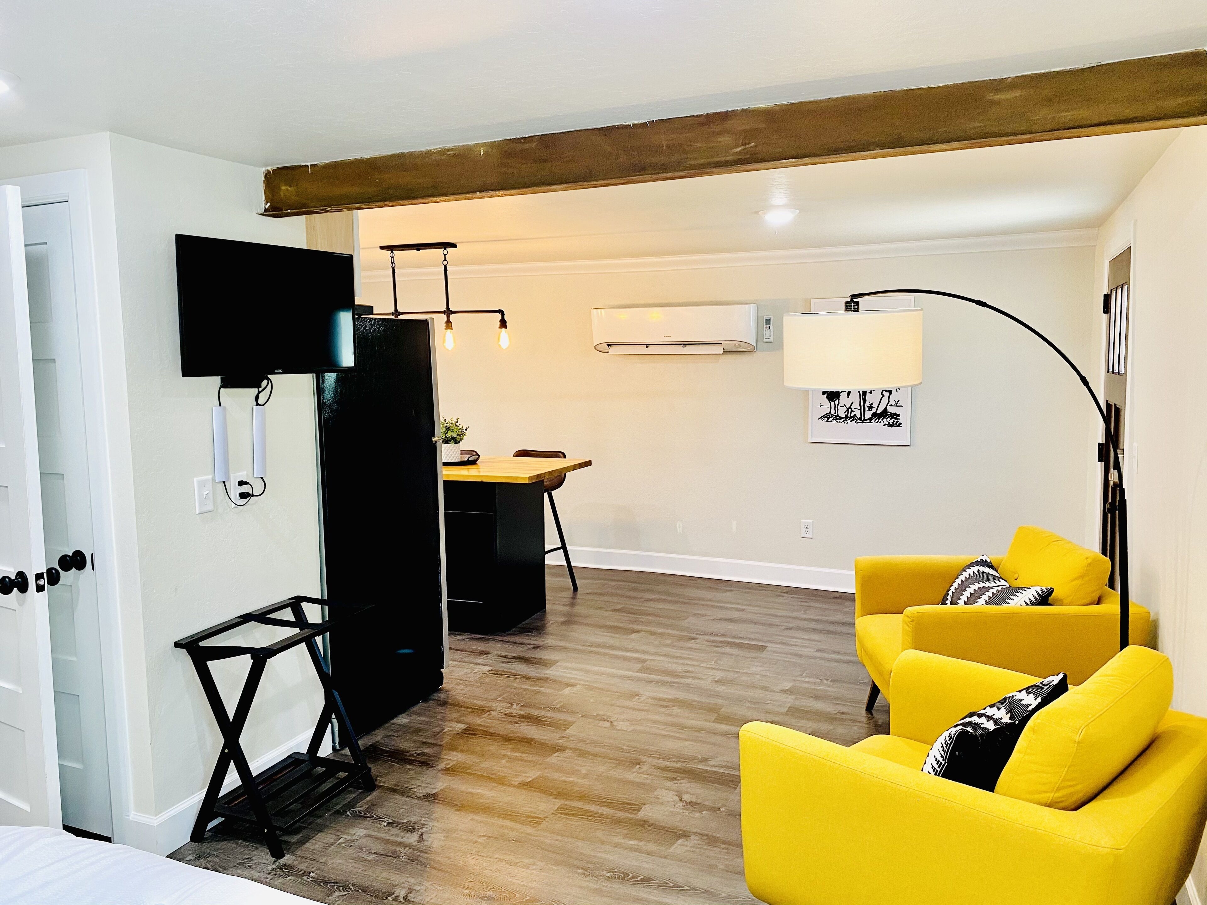 Un salon avec un plancher en bois, un canapé jaune, un meuble de rangement noir pour appareils électroniques et, au fond, un coin repas.