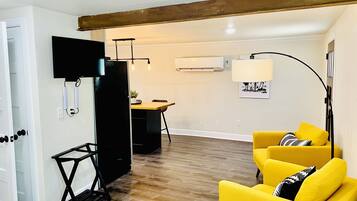 Un salon avec un plancher en bois, un canapé jaune, un meuble de rangement noir pour appareils électroniques et, au fond, un coin repas.