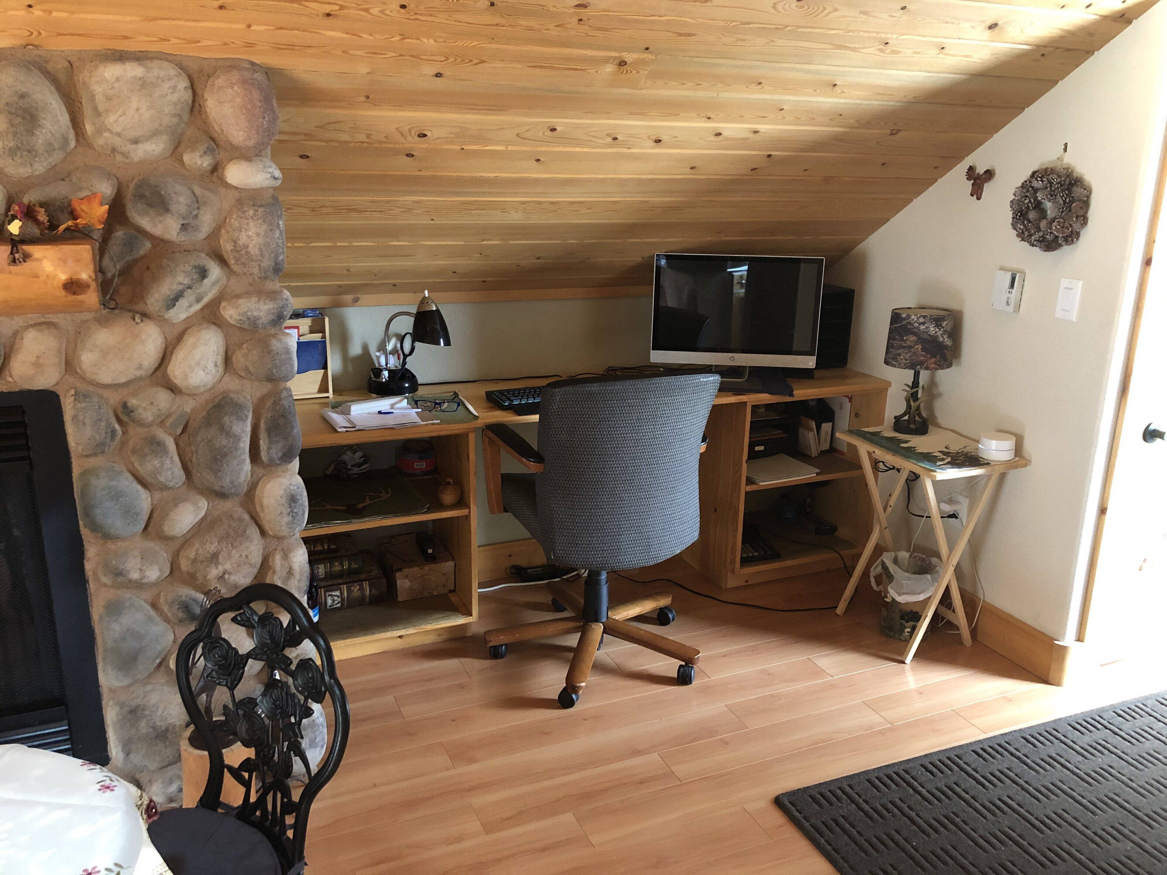 Ein gemütliches Zimmer mit Steinwand, Holzboden und einer Kaminanlage. Es beinhaltet einen Schreibtisch mit Computer, einen Stuhl und weitere Möbel.