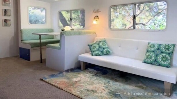 A cozy room with a green sofa, a small table, and a bench. There are windows with tree views and a wall-mounted light fixture. - Cozy Mallard RV. Light, bright and inspiring trailer amongst the Pine trees. (Topanga)