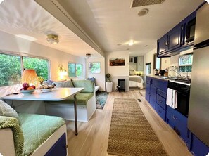 A compact RV interior with a kitchenette, seating area, and a view of greenery outside.