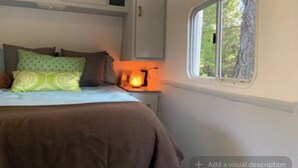 A bedroom with a bed, pillows, a nightstand with a lamp, and a window with a view of trees. - Cozy Mallard RV. Light, bright and inspiring trailer amongst the Pine trees. (Topanga)