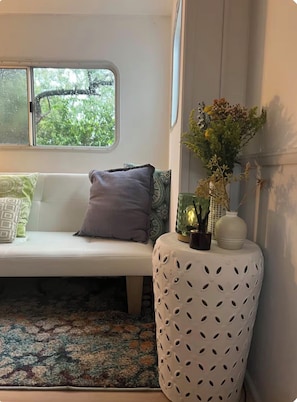 A room with a white sofa, patterned cushions, a decorative vase with flowers, a textured rug, and a window with a view of trees.