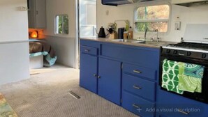 A compact kitchen with blue cabinets, a stove, and a window. - Cozy Mallard RV. Light, bright and inspiring trailer amongst the Pine trees. (Topanga)