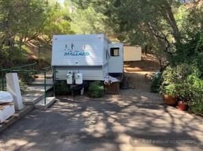 A white RV with 'MALLARD' branding parked in a wooded area with a gravel path, surrounded by trees and potted plants. - Cozy Mallard RV. Light, bright and inspiring trailer amongst the Pine trees. (Topanga)