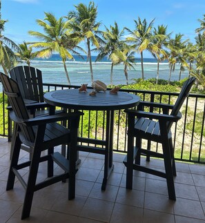 A round table with chairs on a balcony overlooking a beach with palm trees and ocean.