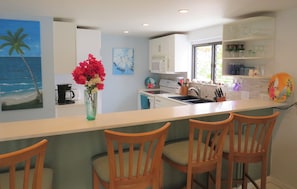Cucina con mobili bianchi, forno a microonde, macchina da caffè e un vaso con fiori sul bancone. Si vedono tre sedie di legno e una finestra con vista sul verde.