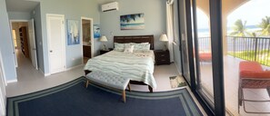 Un dormitorio con cama, mesitas de noche, ventana con vista a la playa y balcón con una silla.