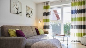 Ein Wohnzimmer mit einer Couch, einem kleinen Tisch und einer Lampe. Vorhänge und ein Fenster mit Blick auf grüne Natur.