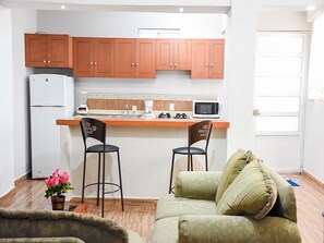 A compact kitchen with wooden cabinets, a refrigerator, a microwave, and a coffee maker. There are two chairs and a sofa with a decorative pillow. - Hospedaje Casa de la Campana Rotamundos (Guanajuato)