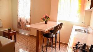A compact kitchen with a dining area, a small table with chairs, a potted plant, and a window with curtains. - Hospedaje Casa de la Campana Rotamundos (Guanajuato)