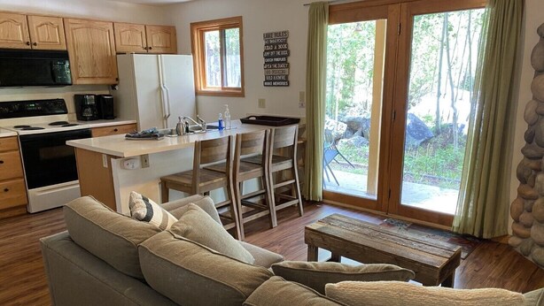 A cozy living room with a sofa, a coffee table, and a dining area with chairs. A kitchen with wooden cabinets and appliances is visible in the background. - Riverfront Condo #1w/patio,1blk 2 town,King bed,ground floor, 40min 2 WinterPark (GRAND LAKE)