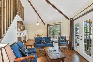 A spacious living area with a wooden staircase, a dining table, blue sofas, and a view of the outdoors through large windows.