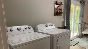 A laundry room with two white Whirlpool washers and dryers, a wooden shelf above the dryers, and a view of a dining area with a chair and table through a window. - 2Bedroom, 1.5Bath Cottage at Beautiful Wood Creek Lake,  Near Wood creek Marina. (London)