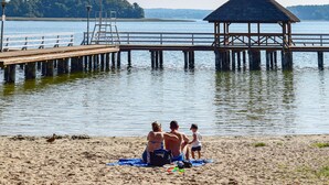 A beach with a wooden pier extending into calm water, a thatched-roof structure, and a sandy area with people and a duck.