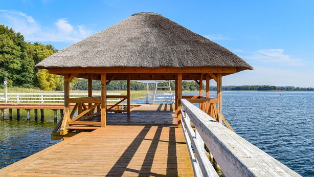 A wooden pier leading to a thatched-roof gazebo by a calm lake.