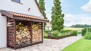 A house with a stone-paved entrance, a wooden pergola with stacked firewood, and a view of a green landscape.