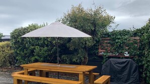 Een houten picknicktafel met bankjes, een parasol en een overdekte barbecue in een achtertuin.