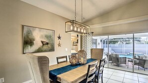A dining area with a wooden table, chairs, a hanging light fixture, and a large window overlooking a backyard with outdoor seating. - We Got You! Loaded home w/ Heated Pool & Large Yard (Fort Myers)