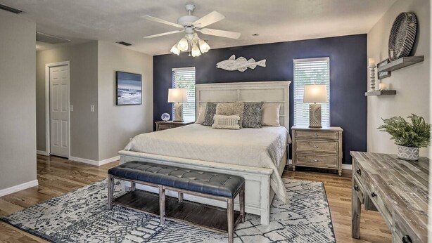 A bedroom with a bed, bedside tables, a bench, a ceiling fan, and a wall-mounted shelf with decorative items. - We Got You! Loaded home w/ Heated Pool & Large Yard (Fort Myers)