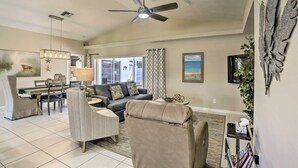 A spacious living room with a ceiling fan, a dining area with a table and chairs, and a large window with a view of the outdoors. - We Got You! Loaded home w/ Heated Pool & Large Yard (Fort Myers)