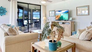 A living room with a sofa, a coffee table with a plant, a TV on a cabinet, and a view of the outdoors through a sliding door. - SS 4224 2/2 Garden View Condo - Paradise Vacation Rental in Ocean Village (Fort Pierce)