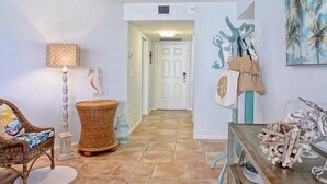 A hallway with a wicker chair, a lamp, a basket, and a table with decorative items. - SS 4224 2/2 Garden View Condo - Paradise Vacation Rental in Ocean Village (Fort Pierce)