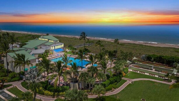 An aerial view of a resort with a swimming pool, palm trees, and a view of the ocean at sunset. - SS 4224 2/2 Garden View Condo - Paradise Vacation Rental in Ocean Village (Fort Pierce)