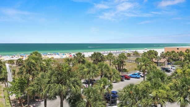 A beach view with palm trees, a parking lot, and a building in the background. - 🌴 Gulf View Condo — Gulf Strand Resort, St. Pete Beach (St Pete Beach)