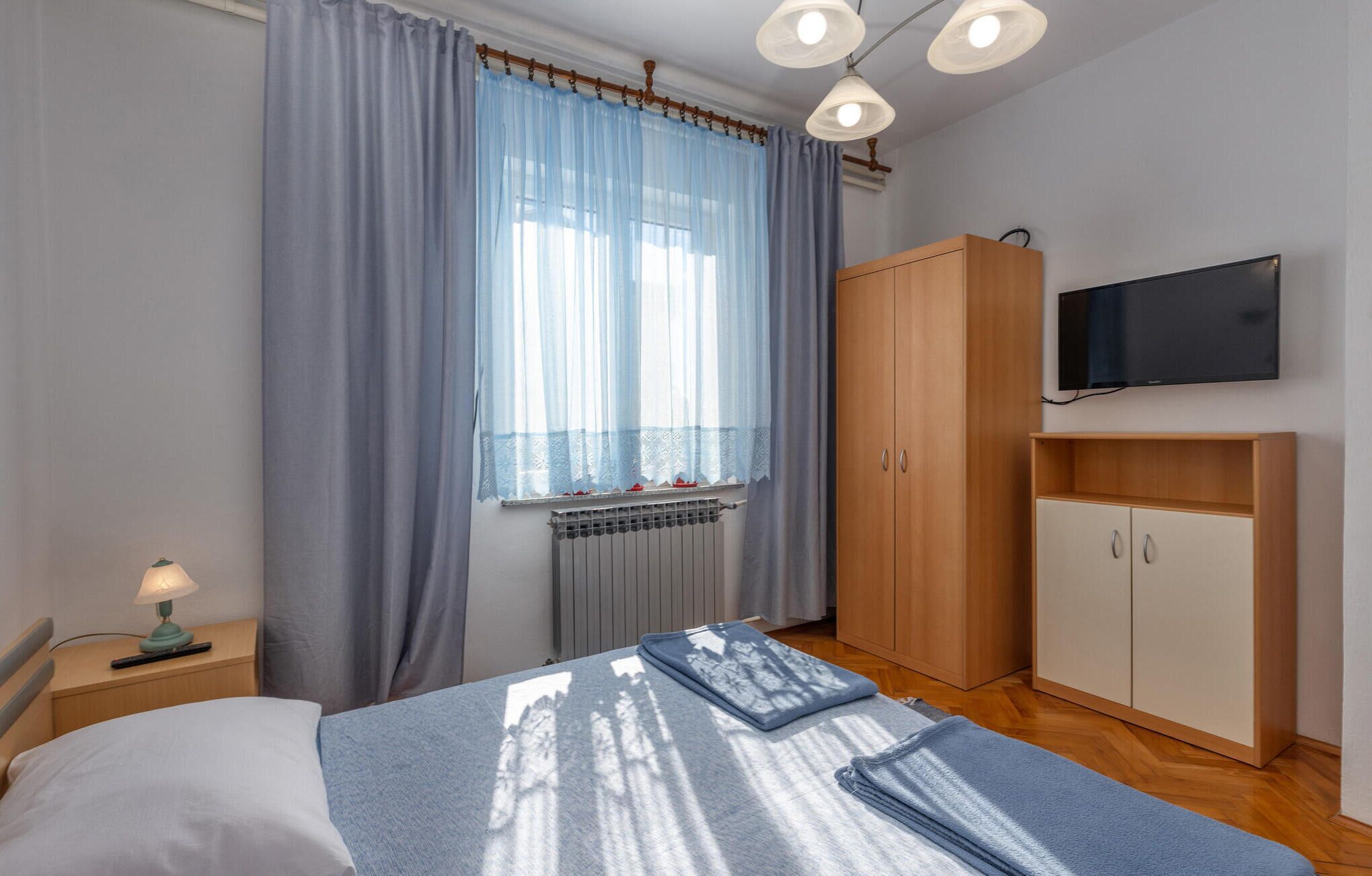Een slaapkamer met een bed, een garderobe, een radiator, een televisie en een raam met gordijnen.