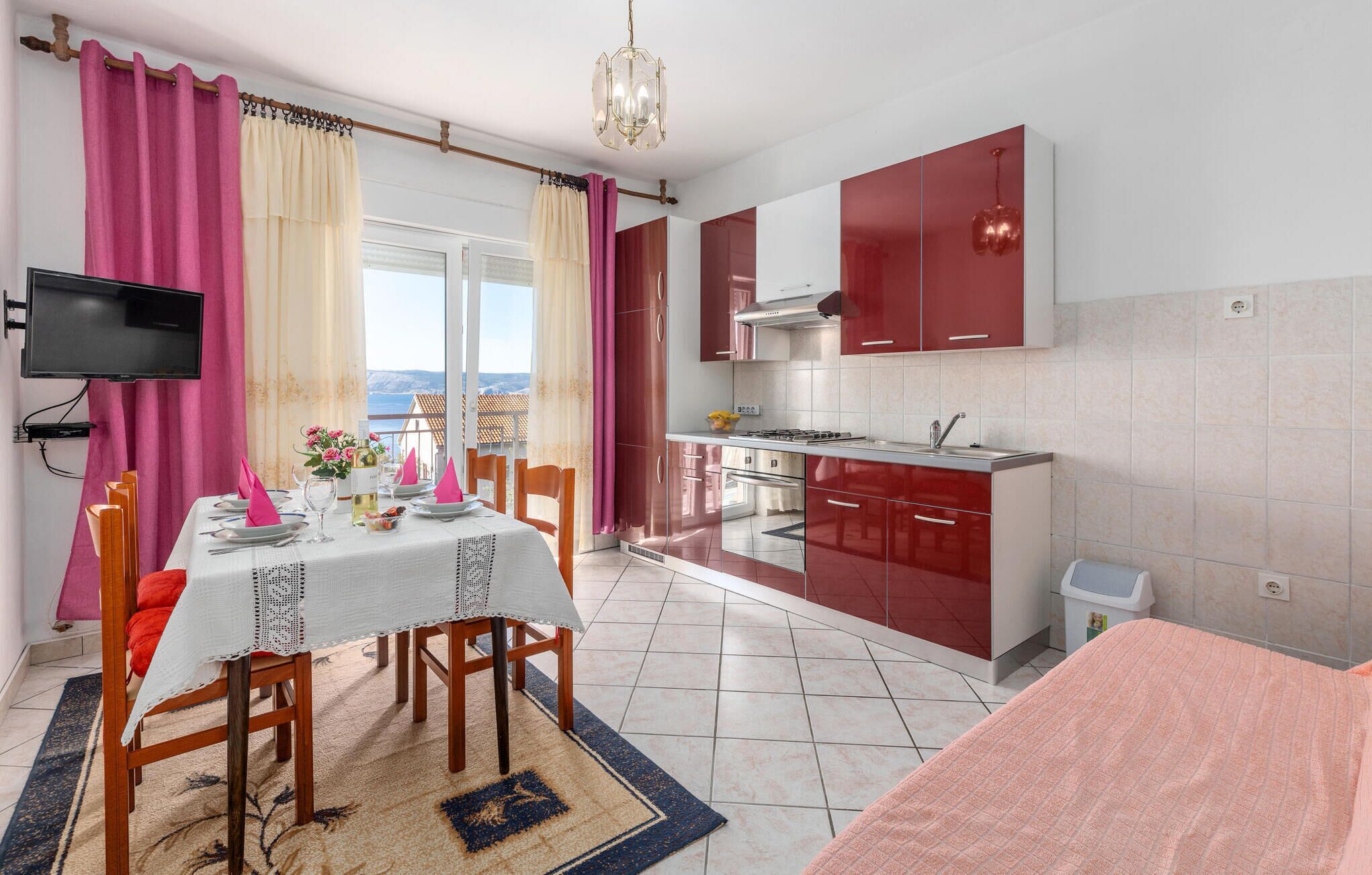 A modern kitchen with red cabinets, a dining area with a table set for two, and a TV mounted on the wall.