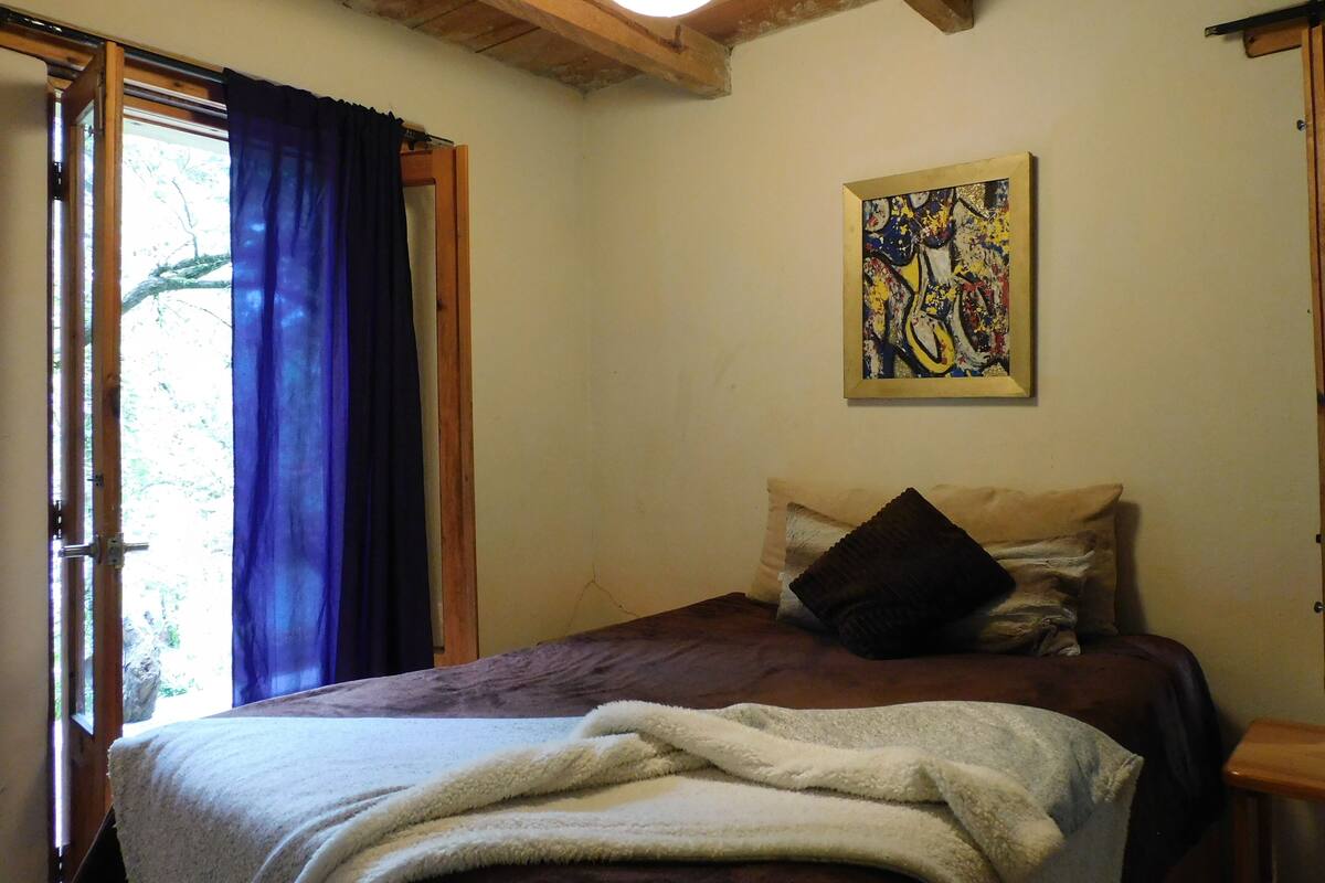A bedroom with a bed, a window with curtains, a painting on the wall, and a wooden ceiling.