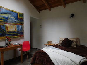 A bedroom with a bed, a desk with a chair, a painting on the wall, and a small table with a plant. - San Nicolás Rotamundos (San Cristóbal de las Casas)