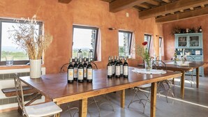 A rustic interior with a long wooden table displaying wine bottles, a vase with dried plants, and a view through large windows.