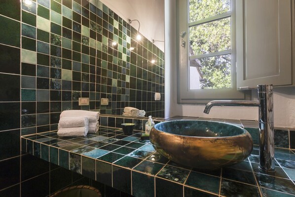 A bathroom with a large copper vessel sink, a towel rack with towels, a small shelf with toiletries, and a window with a view of trees.