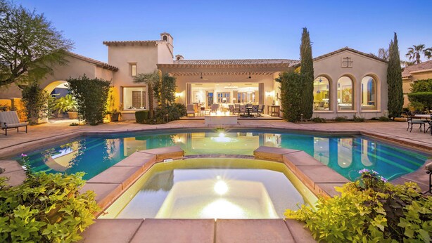 Un área de piscina lujosa con un jacuzzi, rodeada por un jardín cuidado y una casa grande con patio y zona de estar al aire libre.