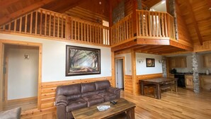 Un intérieur de cabine spacieux, avec un plancher en bois, un canapé en cuir marron, une table basse en bois, une table à manger, un coin cuisine et une mezzanine dotée d’une rambarde en bois.