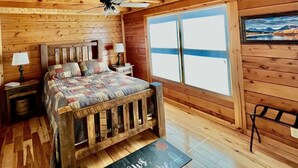 Une chambre confortable aménagée dans un chalet en bois, avec un lit, une table de chevet, une lampe, une chaise et une grande fenêtre donnant sur le paysage extérieur.