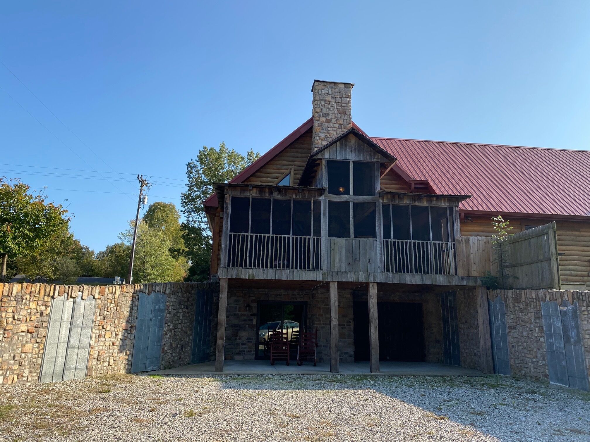 A two-story log cabin with a stone foundation, a red metal roof, and a gravel driveway.